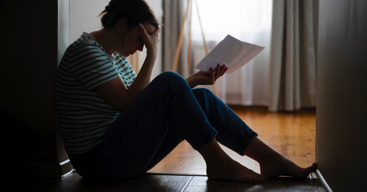 The silhouette of a woman sitting in the hallway of an apartment, rubbing her forehead while holding a piece of paper.