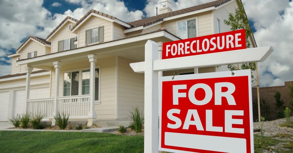 A red and white "FOR SALE" sign with a "FORECLOSURE" notice on top in the front yard of a white house.