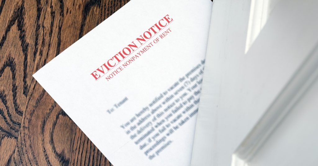 A white paper document with red text "EVICTION NOTICE" slid halfway under a white door on a wooden floor.