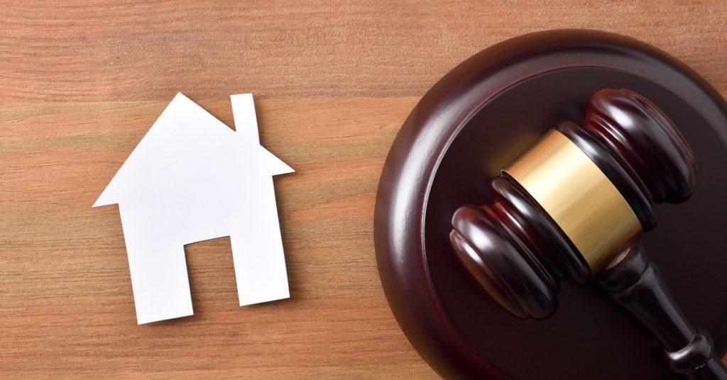 An overhead view of a white paper cut-out of a home next to a courtroom gavel and stand on a wooden table.